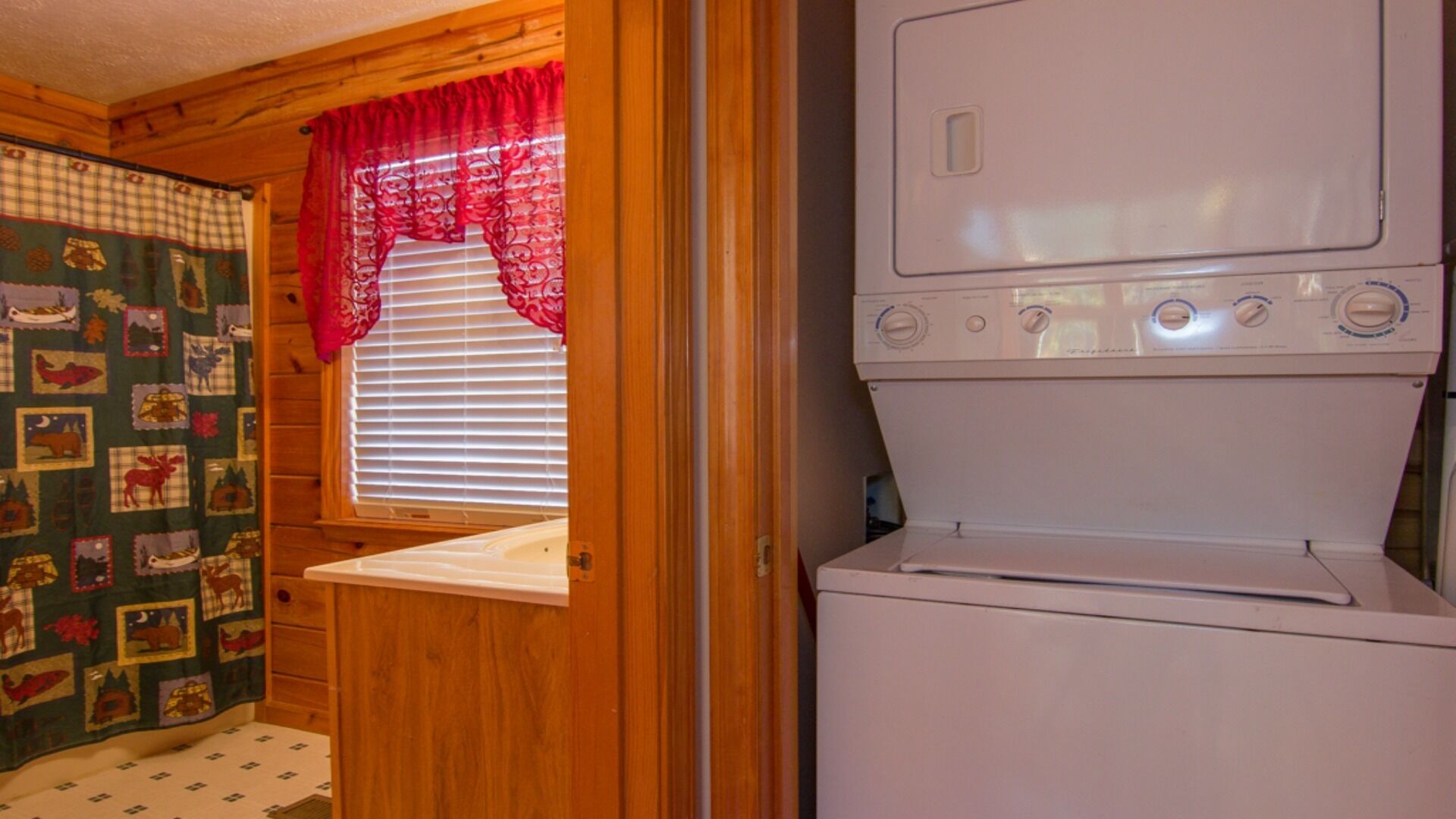 A laundry room with a washing machine and dryer, wooden walls, a window with blinds, and a quilted curtain.