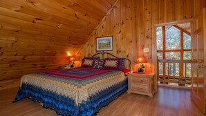 Ein gemütliches Schlafzimmer mit Holzbalkendecke, einem großen Bett mit bunter Bettdecke, zwei Nachttischen mit Lampen und einem großen Fenster mit Blick auf Bäume.