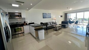 A modern kitchen with a microwave, stove, and oven, a dining area with a table and chairs, a living room with a sofa and coffee table, and a balcony with a view.