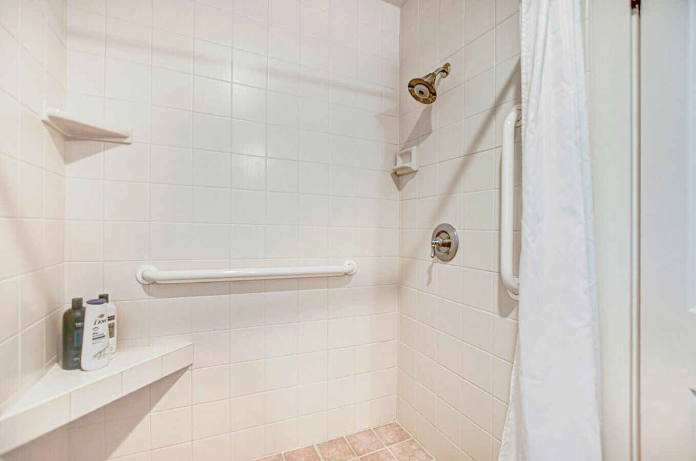 Une salle de bain carrelée de carreaux blancs, une douche avec un pommeau de douche amovible, une barre d'appui et une étagère contenant des articles de toilette.