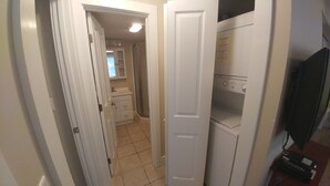 Un couloir étroit avec une porte blanche donnant sur une salle de bain équipée d’un lavabo et d’un miroir.