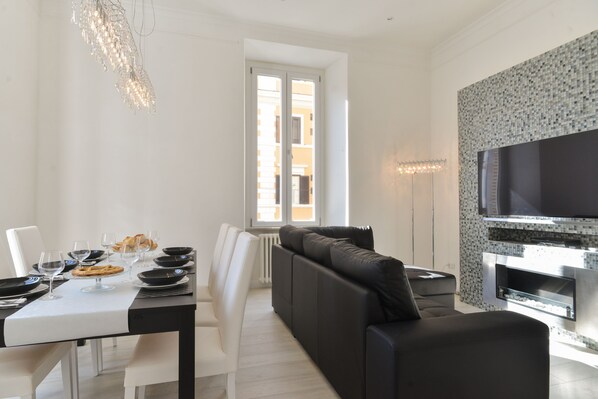 A modern living room with a black sofa, a dining table set for two, and a flat-screen TV mounted on the wall. - Luxury Apt 3 Bedrooms 4 Bathrooms (Roma)