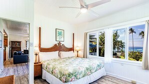 Un dormitorio con cama de madera, ventilador de techo, ventana grande con vistas a palmeras y al océano, y un pequeño rincón de descanso con una silla y una mesa.
