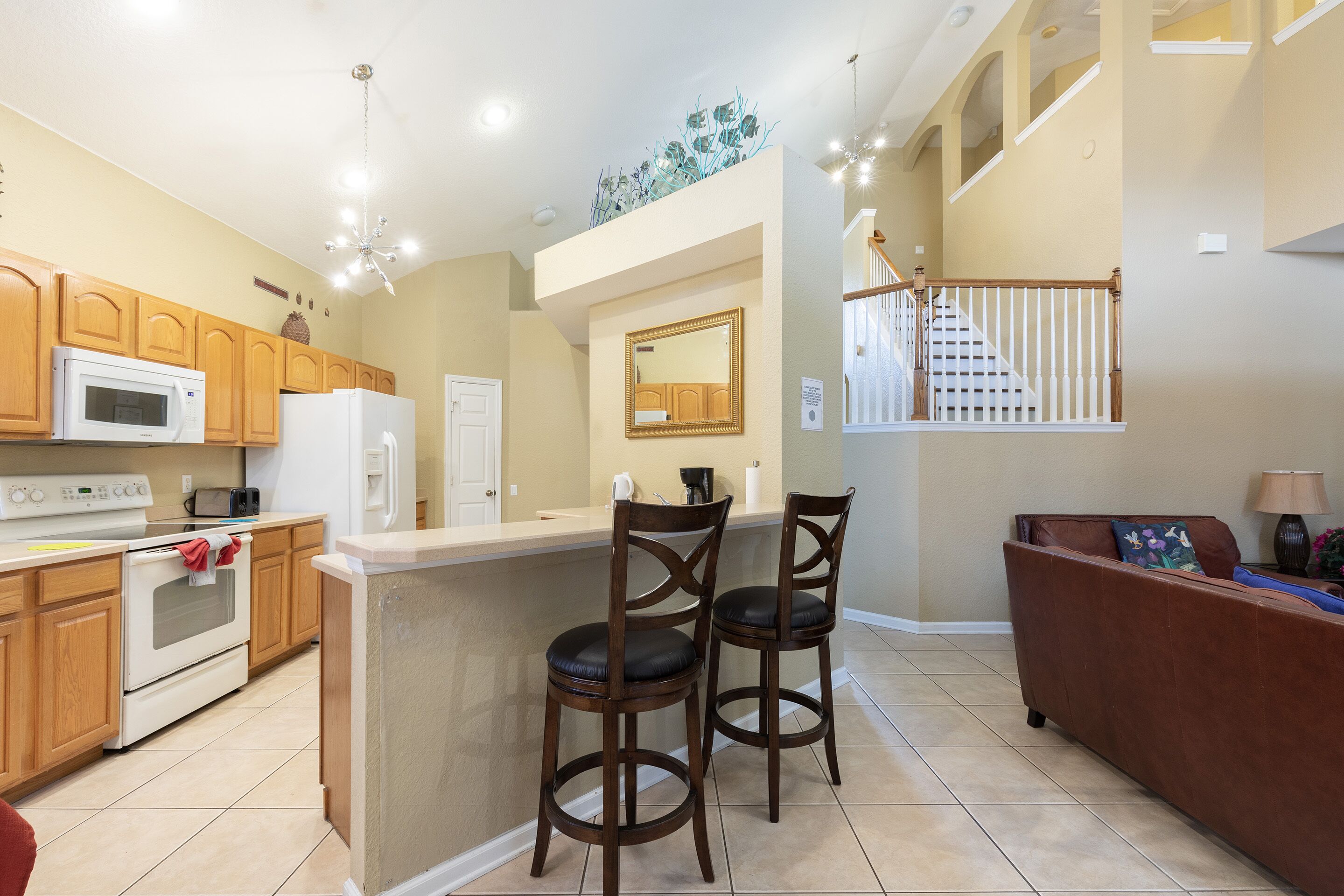 A kitchen with wooden cabinets, a white refrigerator, and a microwave. There are two bar stools with dark seats. A brown sofa is against the wall, and a lamp is on a side table.
