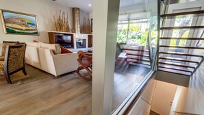 Een moderne woonkamer met een bank, stoelen en een televisie. Er is een trap met een glazen balustrade die naar een hoger gelegen verdieping leidt.
