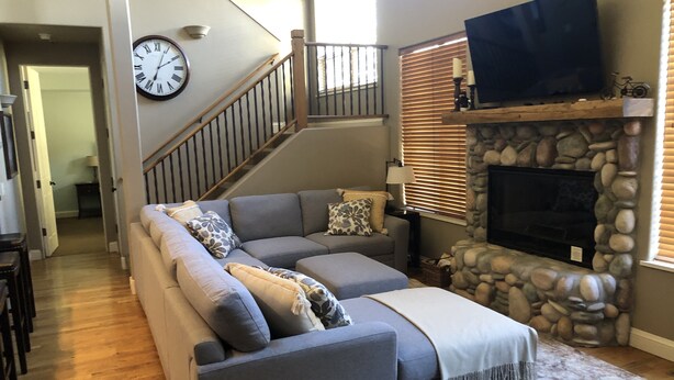 A living room with a gray sectional sofa, a stone fireplace, a mounted TV, and a staircase.