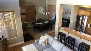 A modern living room with a grey sectional sofa, a dining area with a wooden table and chairs, and a kitchen with a stainless steel refrigerator and dark cabinetry.