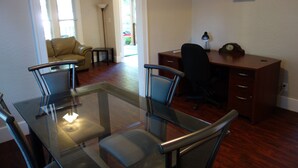 A dining area with a glass table and four chairs, a wooden desk with a chair, and a small shelf with a plant.