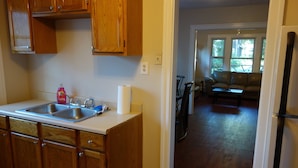 A kitchen with wooden cabinets, a double sink, and a countertop. There is a bicycle in the background, and a living area with a couch and a dining table.