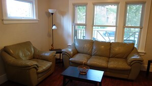 A living room with a brown leather sofa, a matching armchair, and a small table with a basket. There are three windows with blinds, a floor lamp, and a view of the outside.