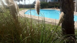 En swimmingpool omgiven av högt gräs och palmer, med en hög byggnad synlig i bakgrunden.