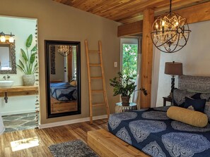 A bedroom with a wooden ceiling, a large bed with patterned bedding, a bedside table with a lamp, a potted plant, and a view of the outdoors through a window.