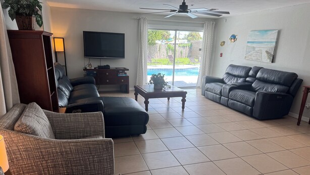 A living room with a flat-screen TV, a ceiling fan, a pool view, and a reclining sofa.