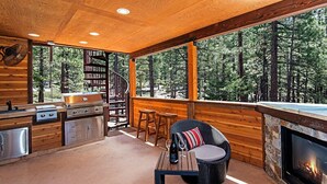 A cozy cabin interior with a wooden interior, a stone fireplace, a grill, and a large window overlooking a forest.