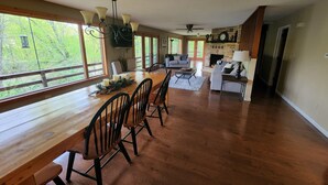 A spacious living room with a wooden dining table, multiple chairs, a fireplace, and large windows offering a view of greenery outside. - Private Mohican Riverfront - Popular Location/Loved by Guests (Loudonville)
