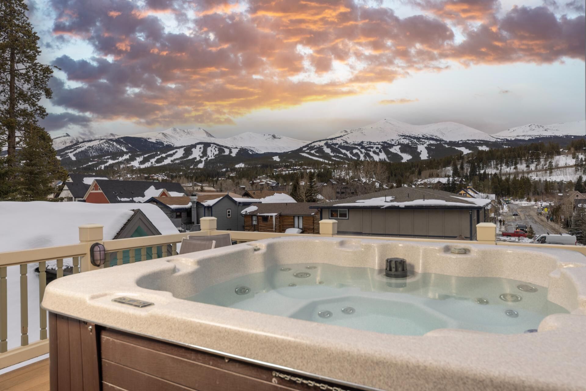 A hot tub on a balcony overlooking a snowy mountain landscape with houses and a sunset.