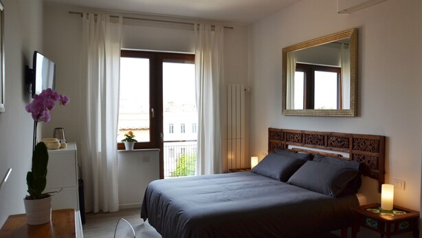 A bedroom with a bed, a TV, a potted plant, a window with curtains, a bedside table with a lamp, and a mirror on the wall. - STUDIO WITH VIEW NEAR THE COLOSSEUM (Roma)