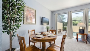 Un coin repas avec une table ronde en bois, six chaises, une plante en pot, un tableau encadré, un téléviseur et une vue sur une piscine, visible à travers de grandes fenêtres.