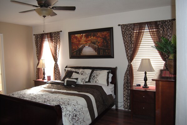 A bedroom with a bed, a ceiling fan, a nightstand with a lamp, and a framed picture on the wall.