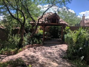 Un espacio de estar al aire libre cubierto, con una estructura de madera rústica, rodeado de exuberante vegetación y un camino empedrado.
