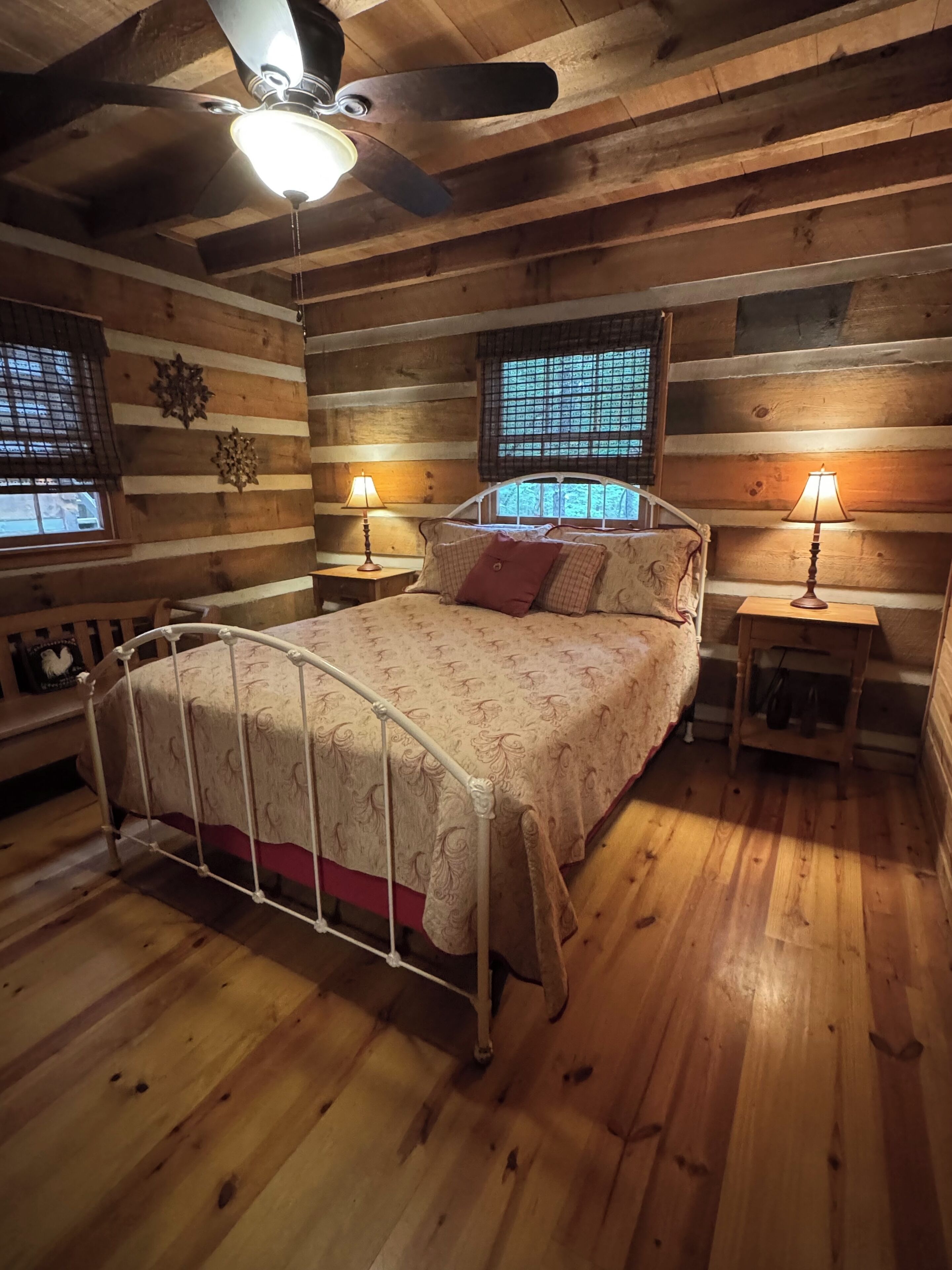 Une chambre confortable dans un chalet en rondins, avec un lit, deux tables de chevet, un ventilateur de plafond et une fenêtre donnant sur un paysage agréable.