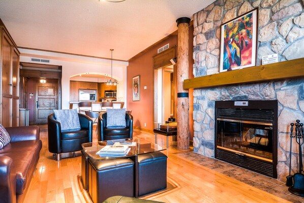 A spacious living room with leather furniture, a stone fireplace, and a kitchen in the background.
