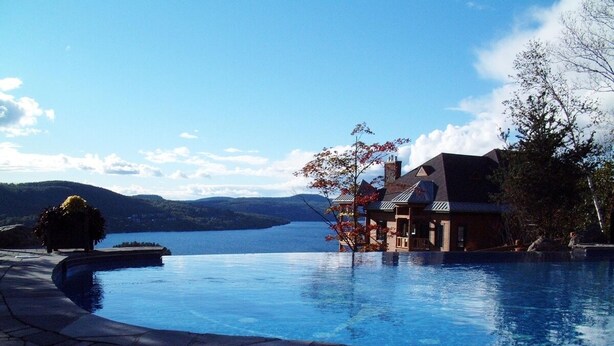 Une piscine avec vue sur un lac et des montagnes, une maison avec un toit rouge et un chemin piétonnier dallé.