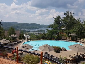Un espace piscine doté de chaises longues et de parasols, entouré d’arbres et offrant une vue sur un lac et des collines.
