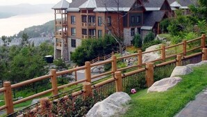 Un grand immeuble à étages, doté d’une façade en pierre et de nombreux balcons, entouré d’une végétation luxuriante et d’une clôture en bois.