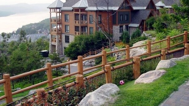 A large, multi-story building with a stone facade and multiple balconies, surrounded by lush greenery and a wooden fence.
