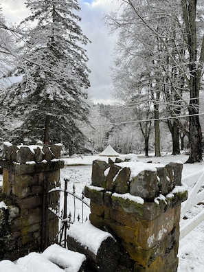 Un muro de piedra con una verja de metal, árboles cubiertos de nieve y una tienda de campaña en un paisaje nevado.