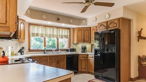 A kitchen with wooden cabinets, a black refrigerator, a window with curtains, and a ceiling fan. - Superb British Colonial holiday home, greenhouse, outdoor deck, tropical garden (Barboursville)