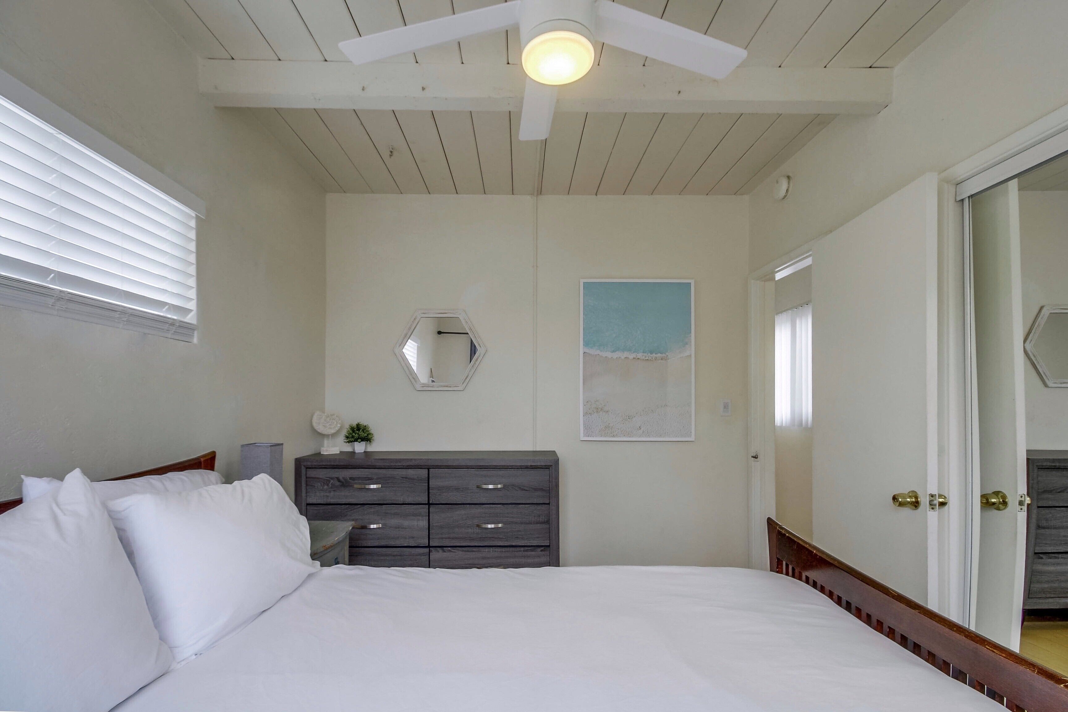 Une chambre à coucher comprenant un lit, une commode, un ventilateur de plafond, une fenêtre avec des stores et une porte.