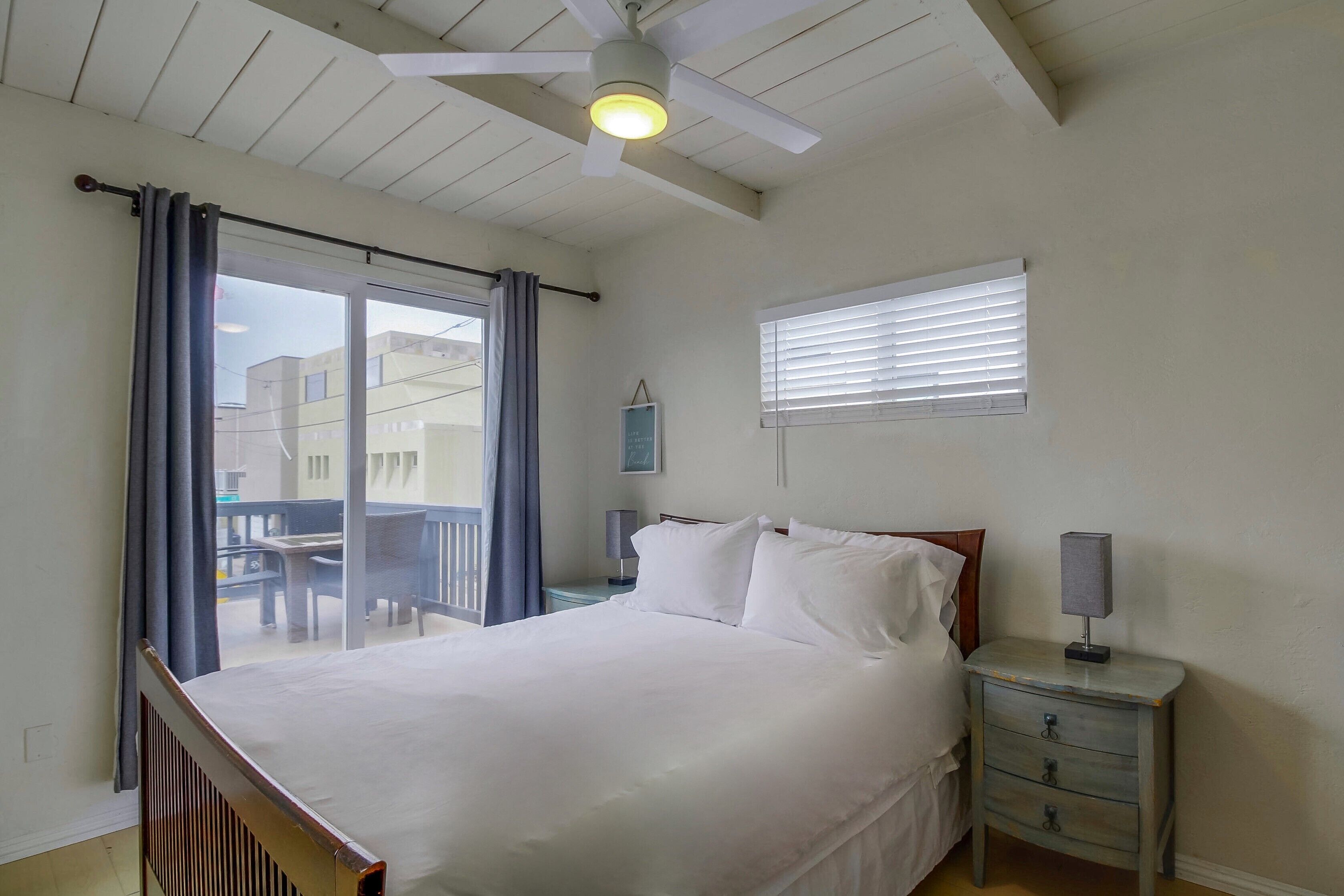 Une chambre à coucher comprenant un lit, une table de chevet, une lampe, un ventilateur de plafond, une fenêtre avec des stores et une vue sur le balcon extérieur.