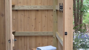 A wooden outdoor sauna with a bench and a towel, surrounded by a wooden fence and a garden with purple flowers. - Great SUMMER WEEKS Available! (New Seabury)