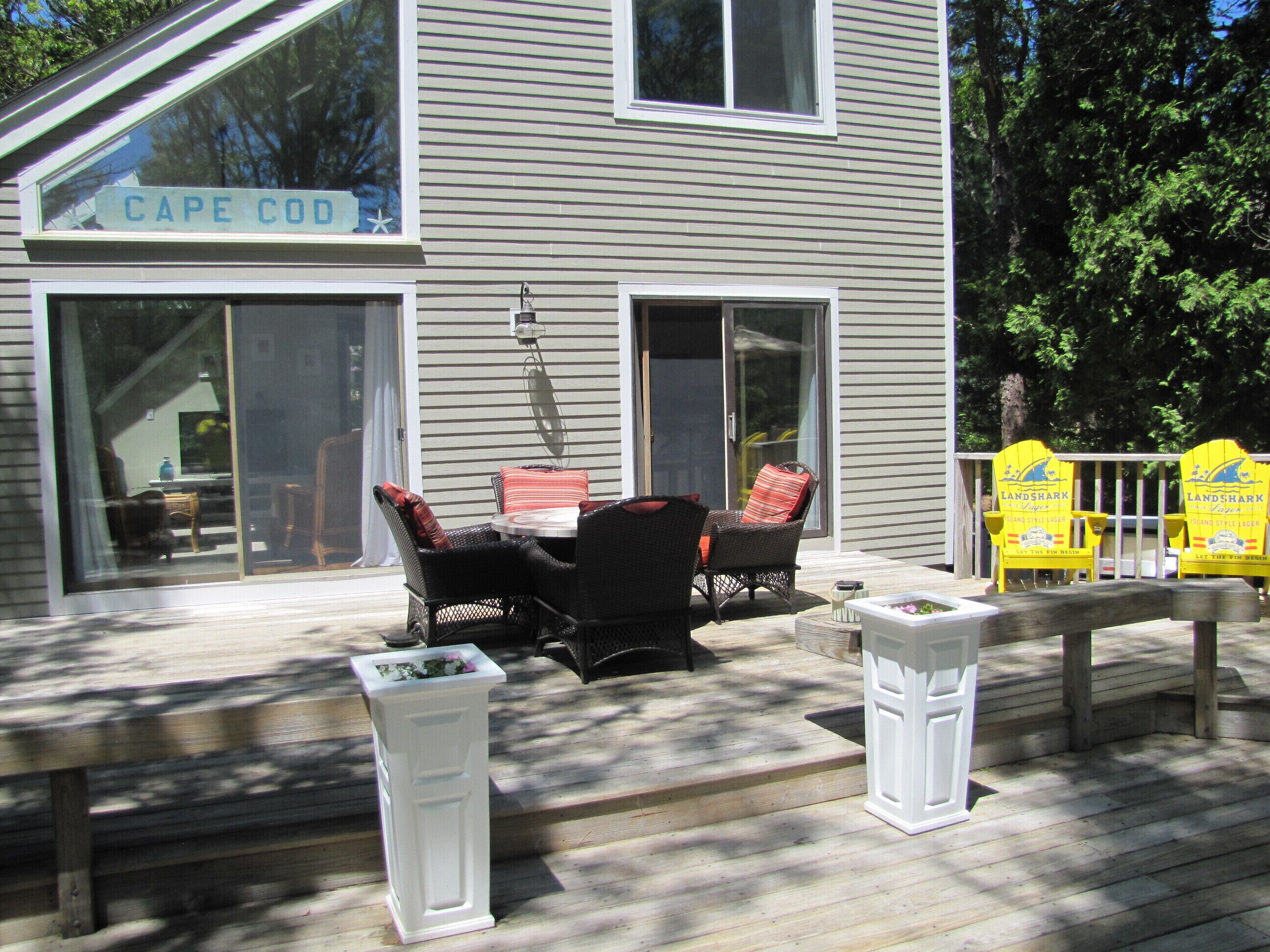 A house with a deck featuring outdoor furniture, a sign reading 'CAPE COD,' and two Adirondack chairs with 'LANDSHARK' branding.
