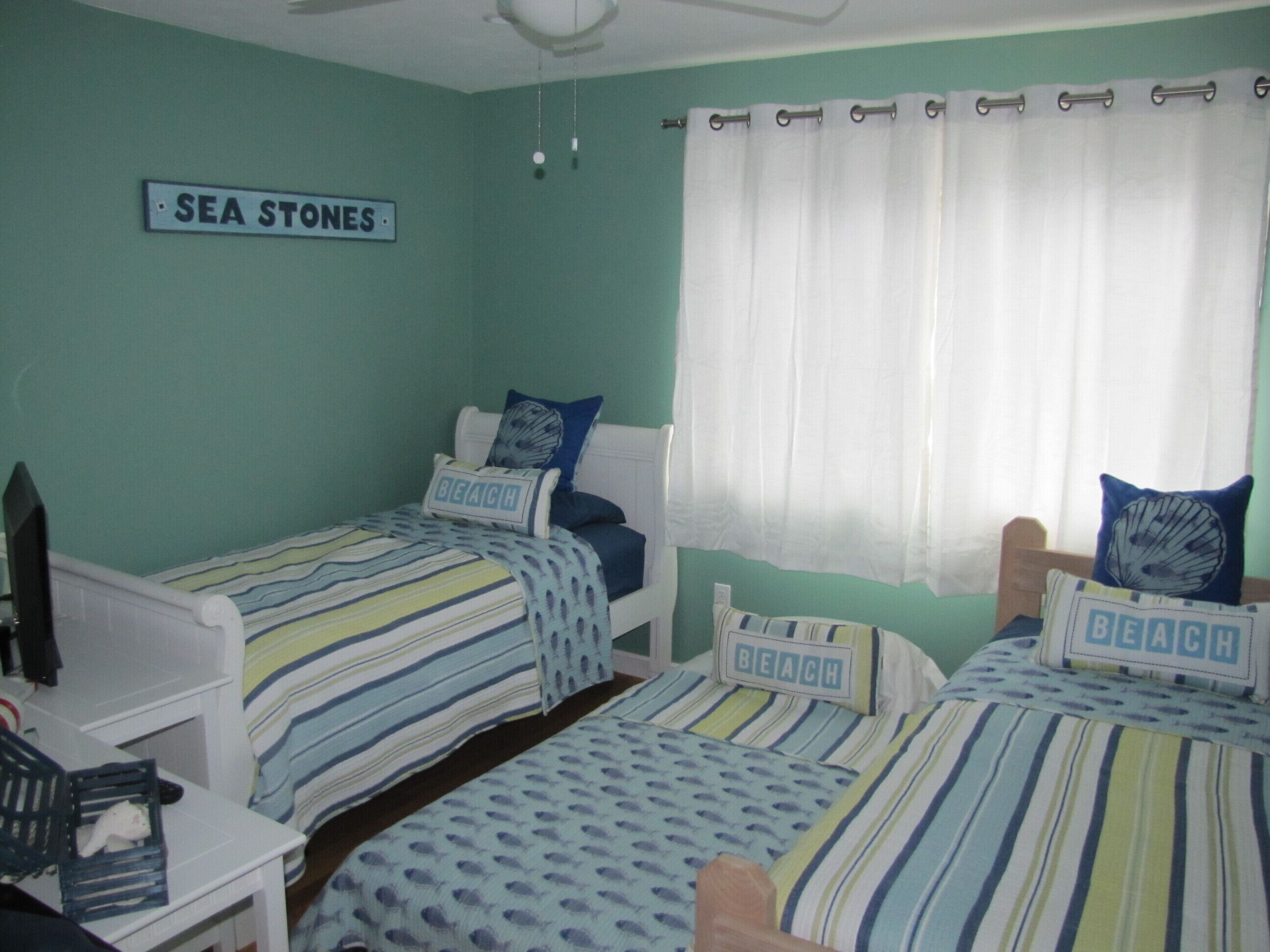 Een kamer met twee eenpersoonsbedden, elk met een gestreept dekbedovertrek en kussens met de tekst 'BEACH'. Een bord op de muur vermeldt 'SEA STONES'.