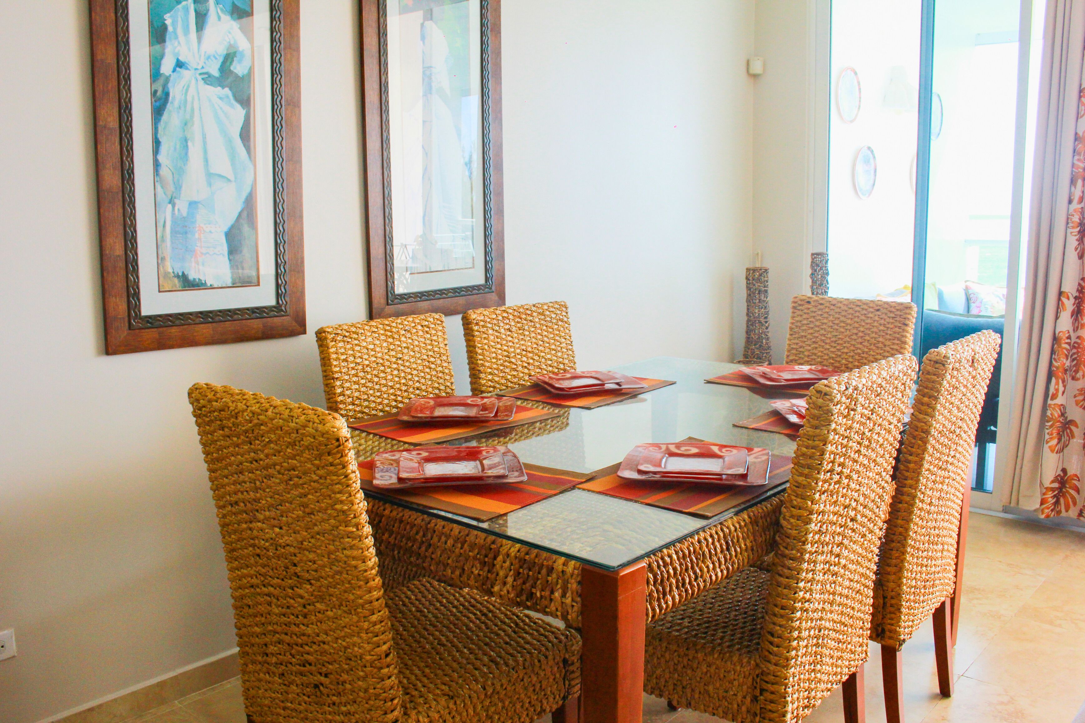 A dining area with wicker chairs and a glass table, framed artwork on the wall, and a view of the outdoors.
