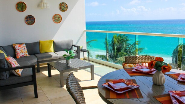 A balcony with a view of the ocean, a sofa, a coffee table, and a dining table set with orange placemats and a vase with flowers. - White Sand Beach Front, Turquois Blue Ocean View, Pools, Hot tub, Guarded,5 star (Christ Church)