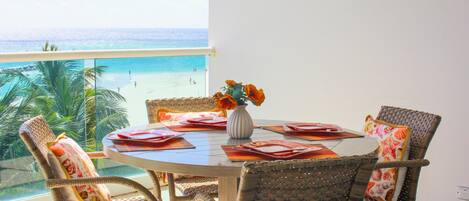 Ein runder Esstisch mit Korbstühlen und einem Blumenstrauß, mit Blick auf einen Strand.