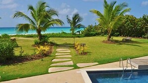 A pool area with a clear view of the ocean, surrounded by palm trees and a well-maintained lawn. - White Sand Beach Front, Turquois Blue Ocean View, Pools, Hot tub, Guarded,5 star (Christ Church)