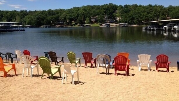 Una hilera de sillas Adirondack coloridas mirando a un lago tranquilo con una orilla boscosa al fondo.