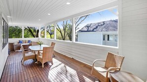 Un patio cubierto con piso de madera, mobiliario de mimbre y ventanas amplias con vistas a árboles y una casa.