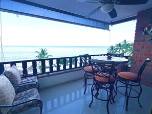 Un balcón con vista al mar, una mesa redonda con sillas y un sofá con cojines estampados.