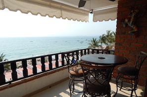 A balcony with a table and chairs, overlooking a beach with boats and palm trees.