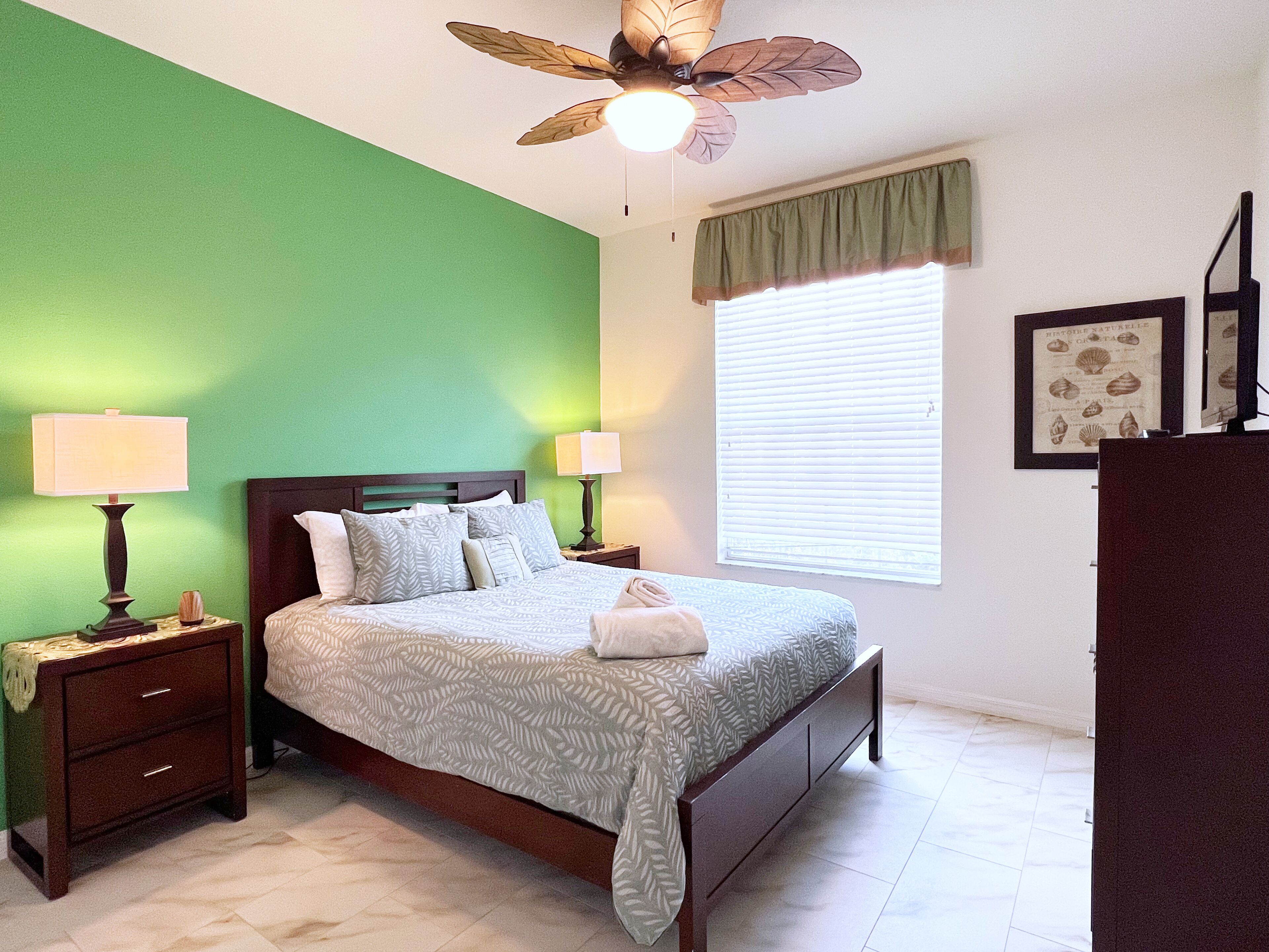 A bedroom with a bed, two bedside tables, a ceiling fan, a window with blinds, and a dresser.