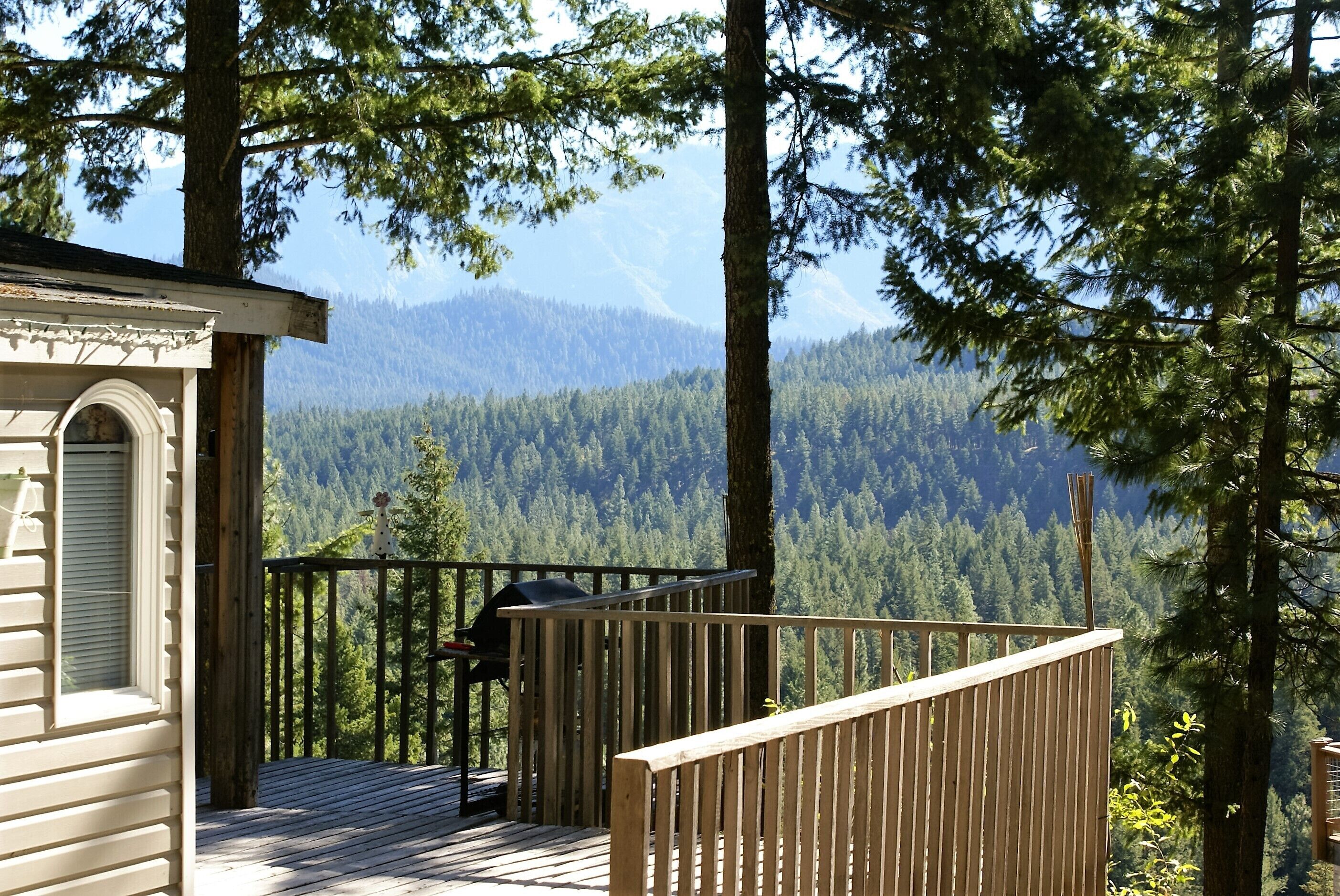 Une terrasse en bois avec un barbecue, un petit bâtiment doté d’une fenêtre, et une vue sur une chaîne de montagnes boisées.