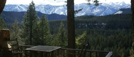 Une terrasse en bois, agrémentée de chaises en métal, qui offre une vue sur une vallée boisée et des montagnes aux sommets enneigés.