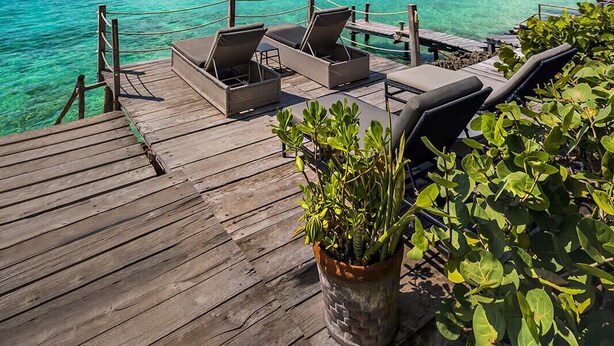 A wooden deck with lounge chairs and a potted plant, overlooking a clear blue sea and a pier.