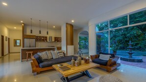 A spacious living area with a sofa, coffee table, and ottomans, a kitchen in the background, and large windows offering a view of the outdoors.